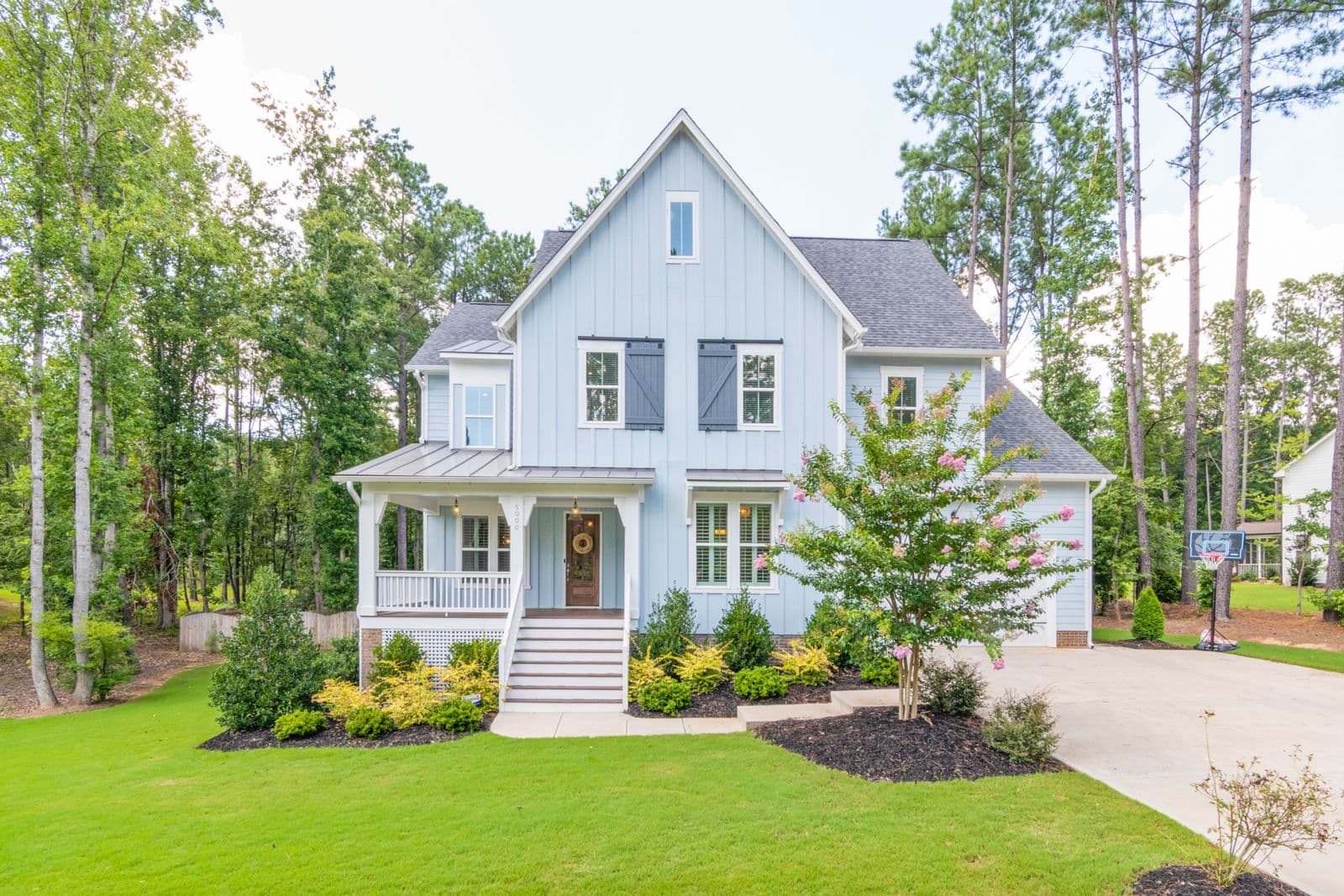 A light blue farmhouse-style home with a covered front porch, framed by tall pines