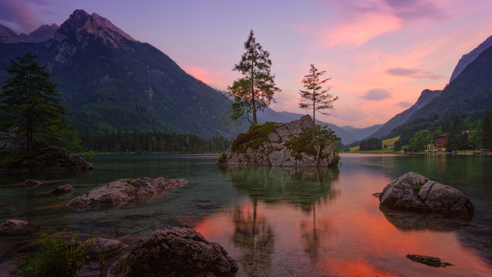 A mountain lake at sunset, rock outcrops and pines reflected in still water lit pink and orange