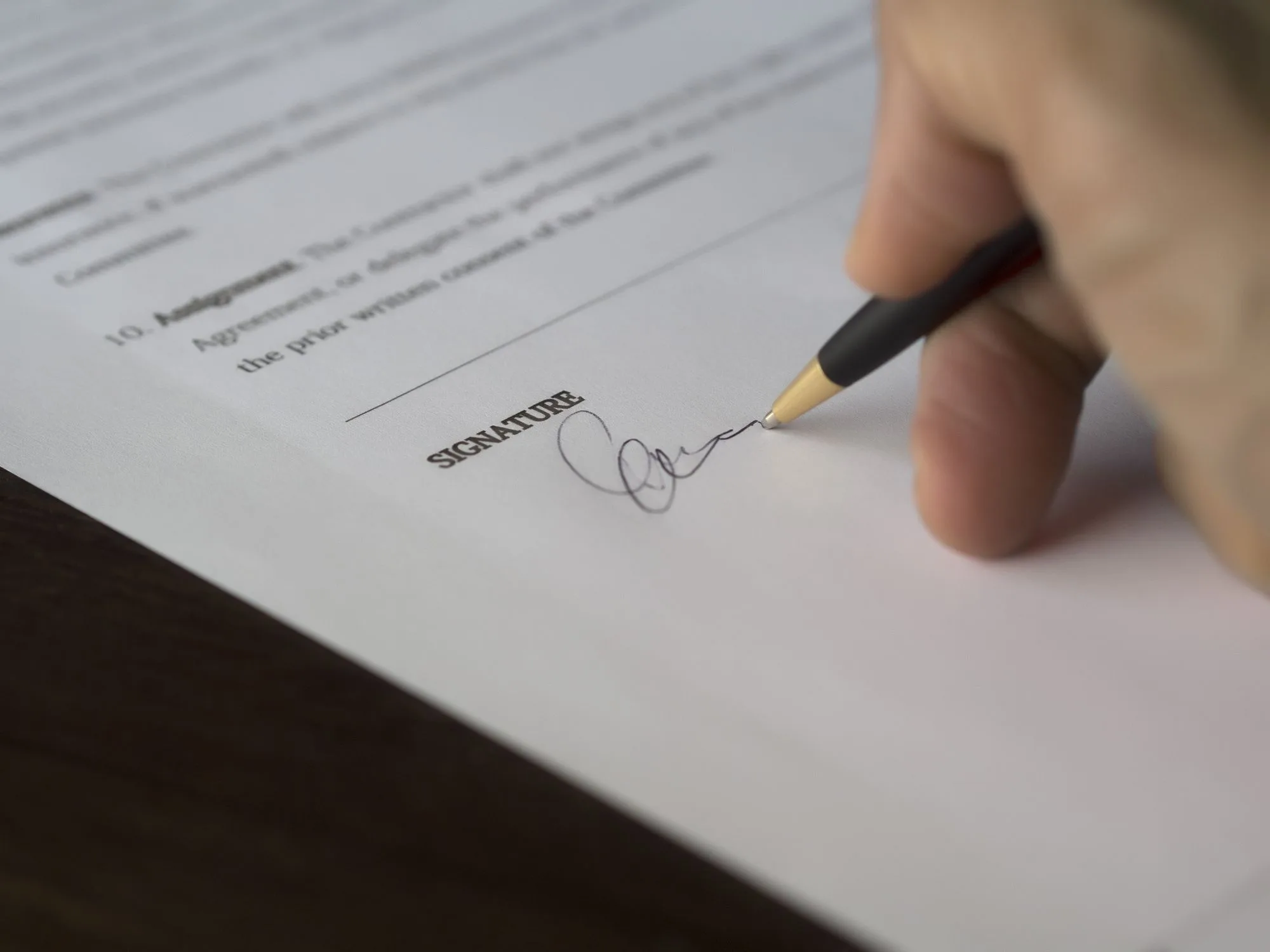 A hand signing a contract with a pen on a dark wooden desk