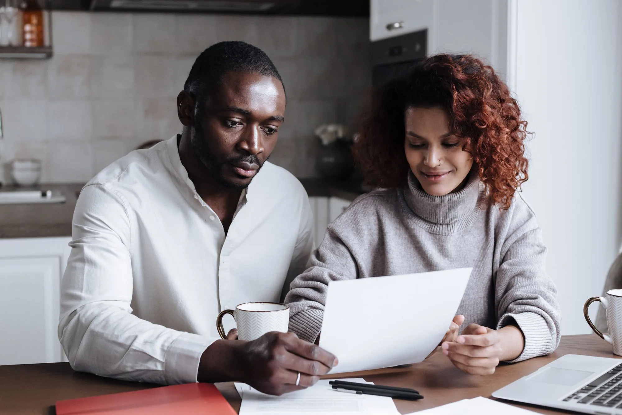 A couple reading a printed page together at a wooden kitchen table with coffee