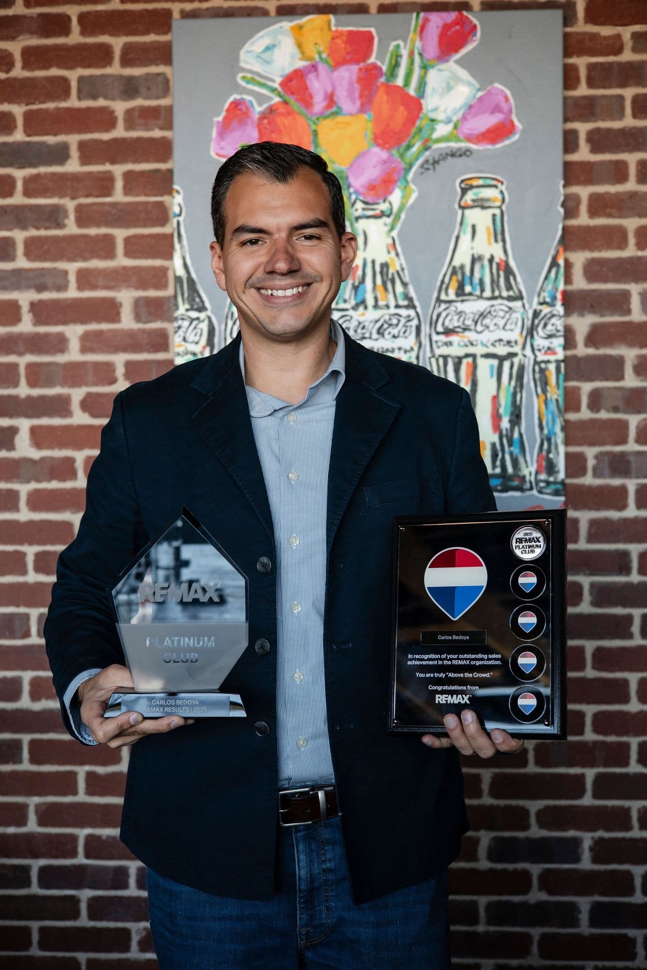 Carlos Bedoya smiling in front of a brick wall, holding a RE/MAX Platinum Club award and plaque
